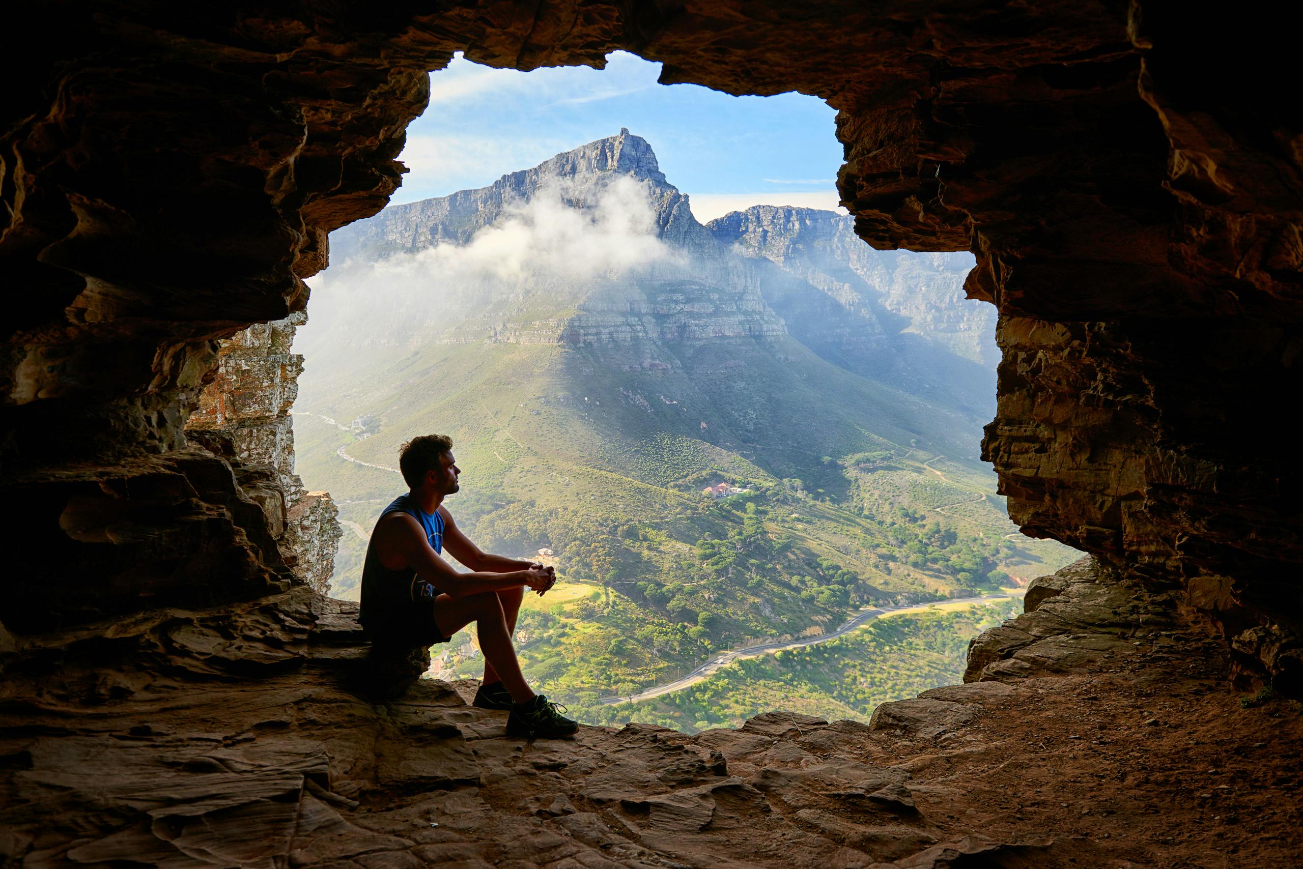 A man sitting in a cave overlooking a majestic mountain landscape under daylight.