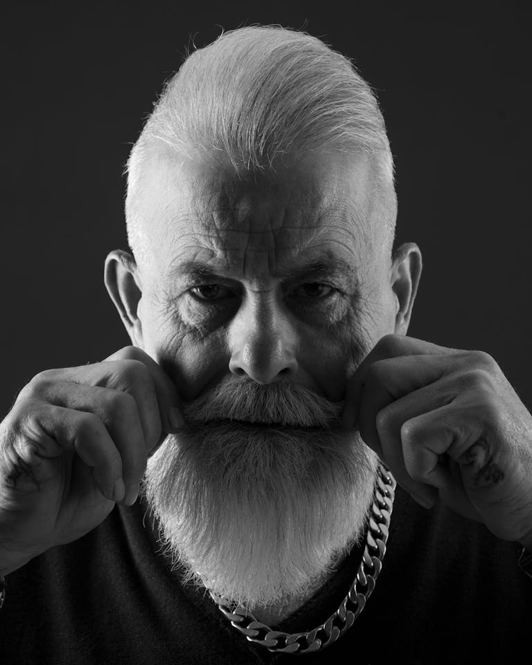 Black and white portrait of a senior man with beard and chain, adjusting facial hair.